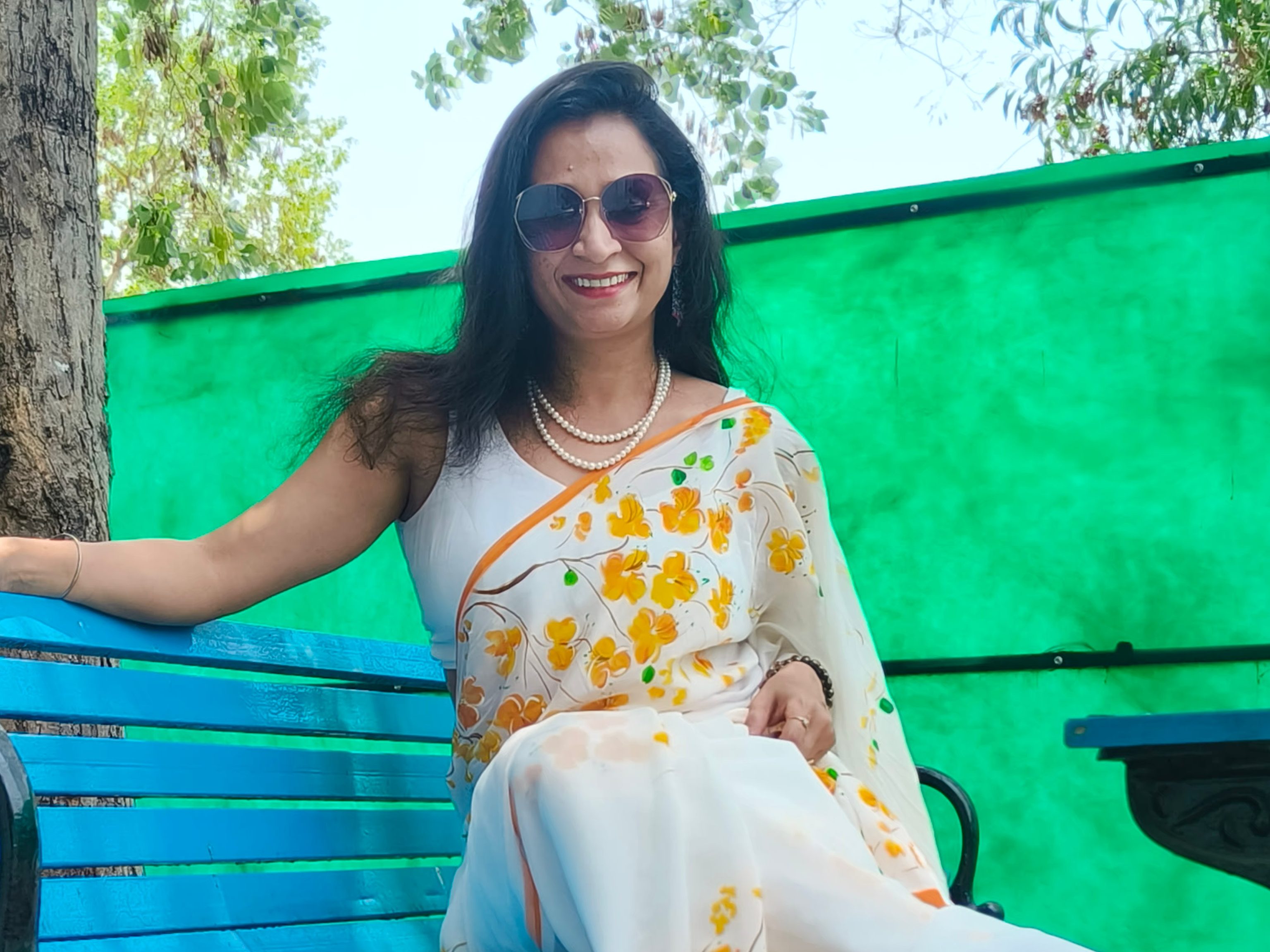 A woman wearing a floral saree sitting on a blue bench under a green tree, smiling at the camera.